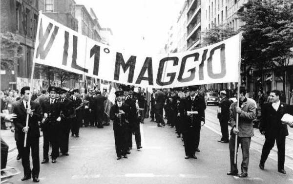 Manifestazione per il primo maggio in Italia, negli anni cinquanta. In testa al corteo ritratto nella foto in bianco e nero, davanti alla banda musicale, due uomini sorreggono uno striscione che recita 'W il 1° maggio'.